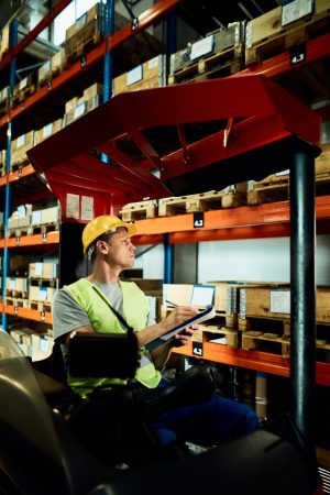 Forklift operator taking notes while examining stock of packages in a warehouse.
